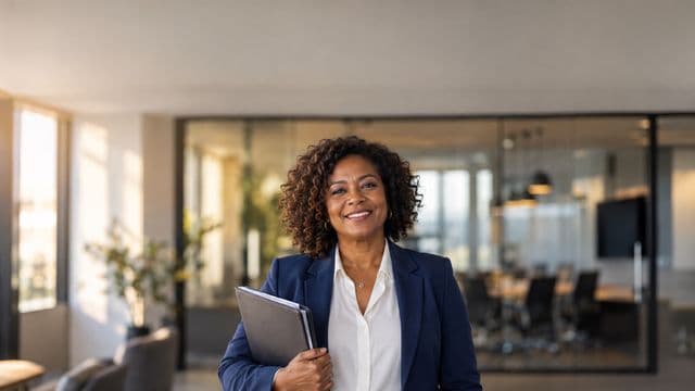 Profissional de recursos humanos sorrindo, com blazer azul sobre camisa branca, segurando uma pasta de documentos, em um escritório moderno com uma sala de reunião envidraçada