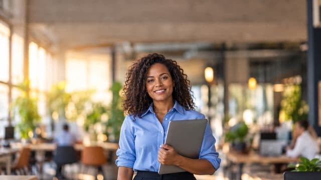 Empreendedora sorrindo, com camisa social azul com as mangas dobradas, segurando um notebook, em um espaço de coworking moderno e iluminado